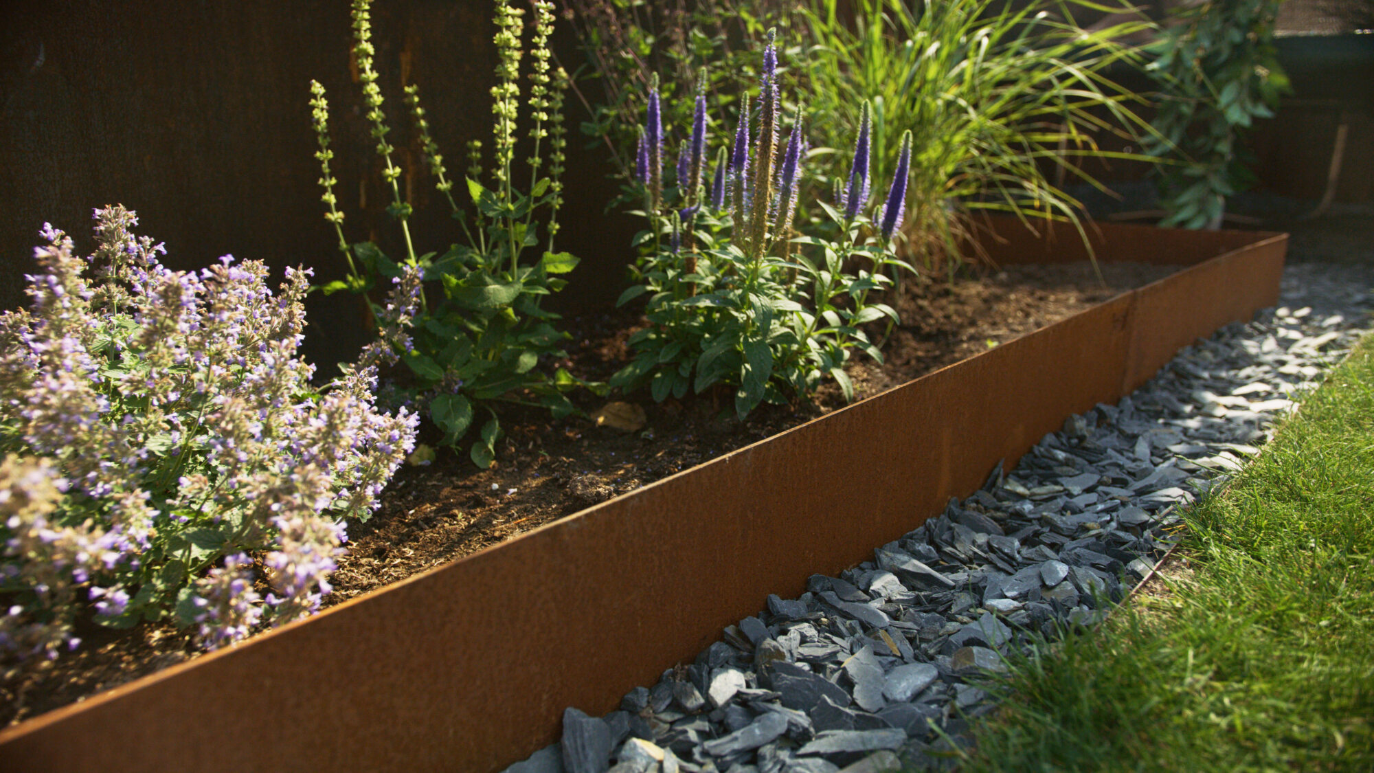 Weathered steel garden edging surrounding a flower bed with purple and green plants, creating a structured and eco-friendly garden space.